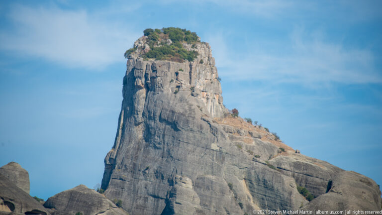 Steep gray sandstone pillar with streaked rock faces and shrubs and small trees growing on its narrow summit.