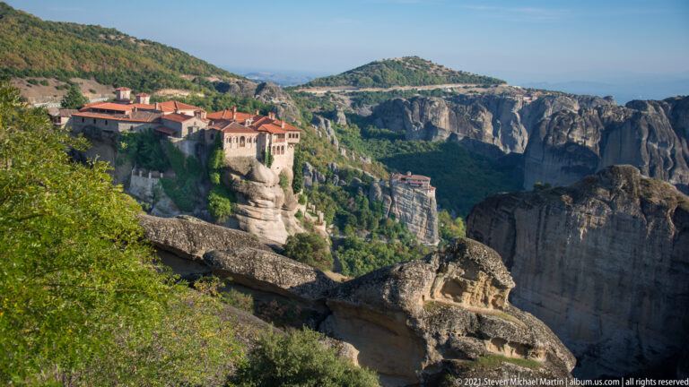 Weathered rock ledges frame a distant monastery complex, with cliffs and another smaller monastery beyond.