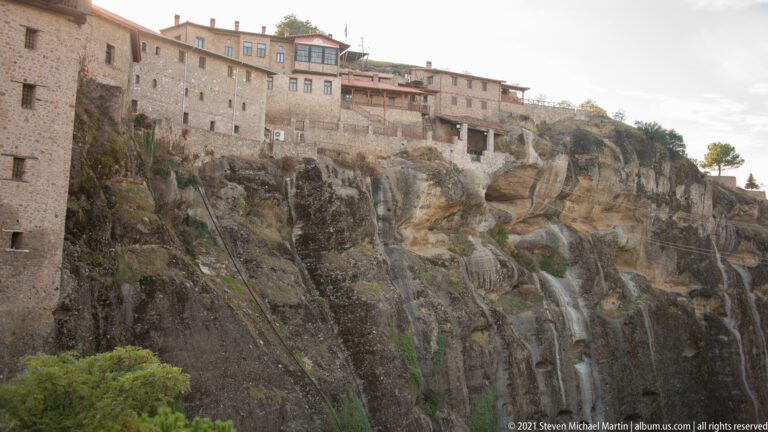 Stone monastery buildings line the top of a steep rocky cliff above a narrow path and retaining walls.