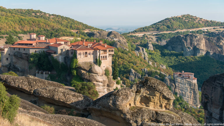 Rounded sandstone ledges lead toward a large monastery, with a second rock-top monastery visible farther right.