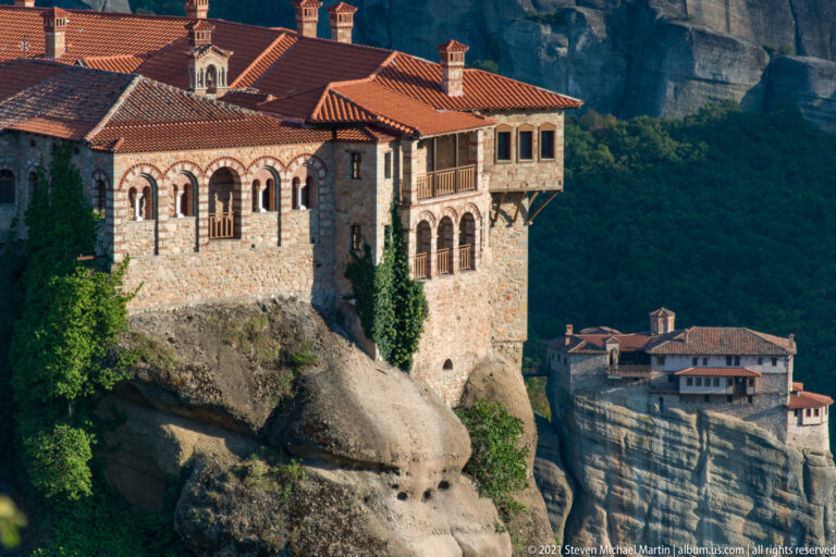 Arcaded stone monastery with red roofs sits on rounded rock, with another monastery visible across the valley.