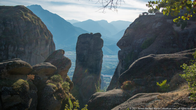 Tall isolated sandstone pinnacle stands between darker rock masses, with hazy mountain ridges in the distance.