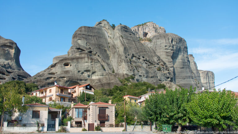 Red-roofed houses and trees at the foot of a huge weathered sandstone cliff under a bright blue sky.