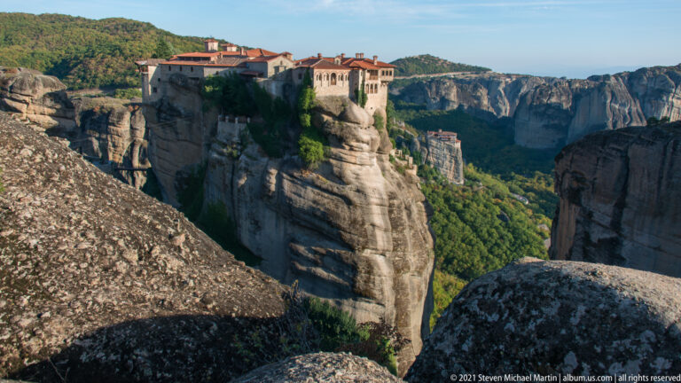 Large weathered boulders frame a monastery across a deep ravine, with another rock-top complex in the distance.