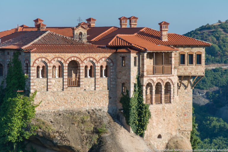 Close stone monastery façade with arched windows, wooden balconies, red tile roof, and several chimney stacks.