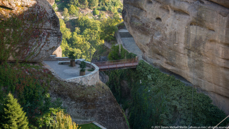 Paved terrace and railed footbridge cross a narrow vegetated gap between massive sandstone walls.