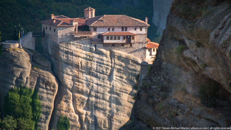 Red-roofed monastery complex sits directly on the edge of a tall, vertically striated sandstone pillar.