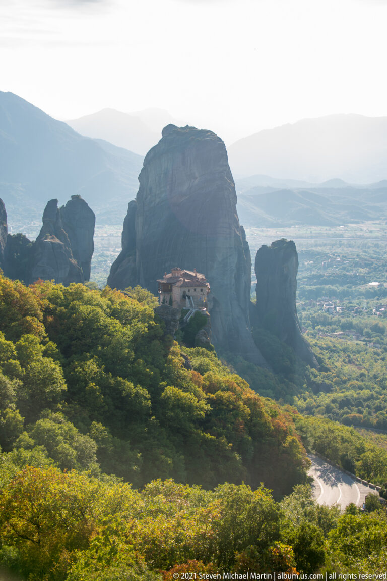 Small monastery sits among green trees below enormous rounded sandstone towers in a hazy mountain landscape.