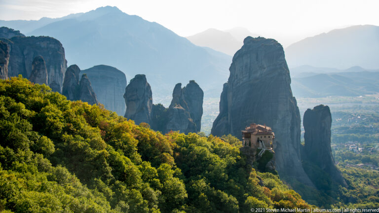 Monastery rises above dense forest at the base of towering sandstone pillars, with hazy mountains beyond.