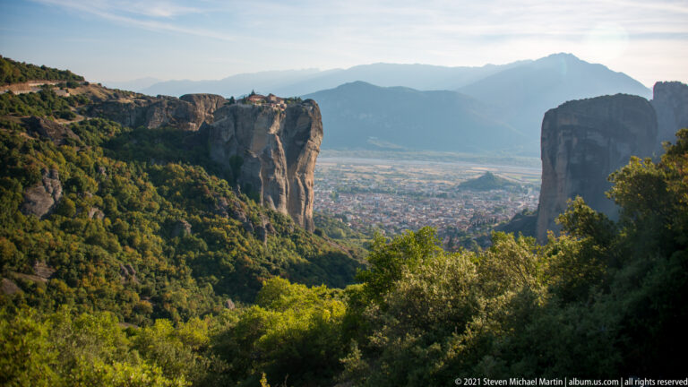 Monastery crowns a distant sandstone cliff above a town-filled valley framed by wooded slopes and rock walls.