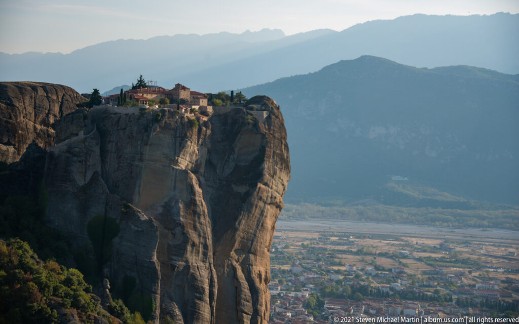 Small monastery crowns an isolated sheer sandstone tower above a broad settled plain and distant mountains.