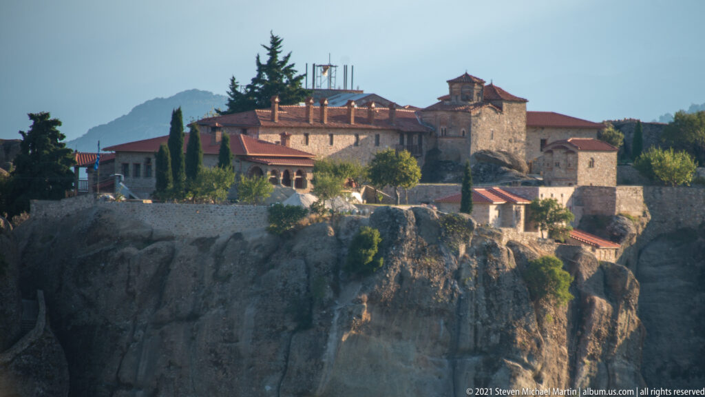 Stone monastery with red tiled roofs, church tower, and cypress trees stands on a sheer sandstone edge.