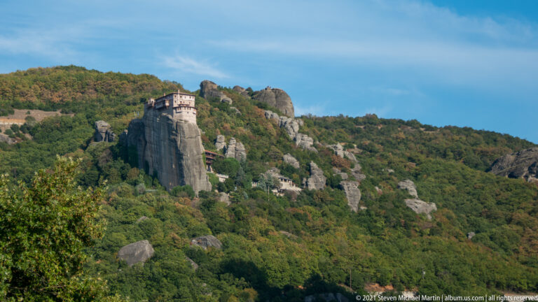 Compact monastery perched on a narrow sandstone pillar above dense green forest and scattered rock outcrops.