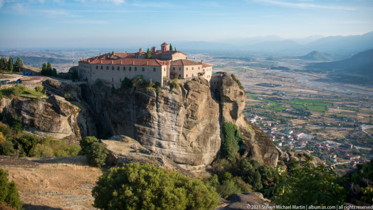 Large pale stone monastery with red roofs stands on a high rock outcrop above fields, town, and distant mountains.