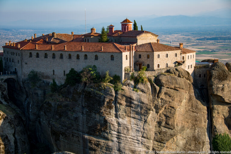 Large stone monastery with red roofs and central church sits directly atop a deeply eroded sandstone cliff.