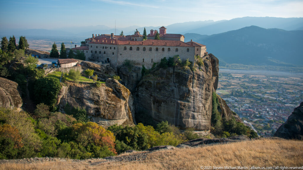 Large monastery crowns a sandstone outcrop beyond dry grass and trees, with plain and mountains behind.