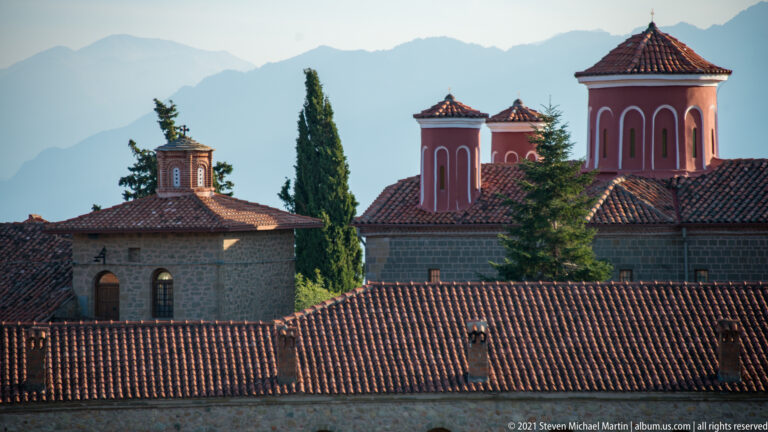 Red tiled roofs fill the foreground below pink church towers and tall cypress trees against distant mountains.