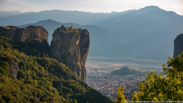Small monastery crowns a narrow sandstone tower above forested slopes, town, plain, and distant mountains.