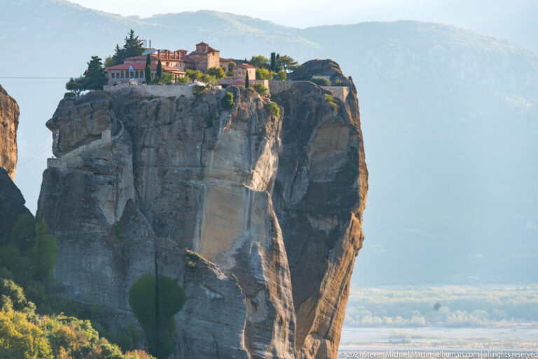 Monastery complex extends across the top of a massive sheer sandstone tower against hazy mountains.