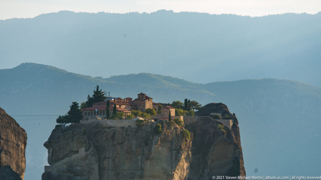 Compact stone monastery and trees sit on a sandstone crest against layered blue-gray mountains.
