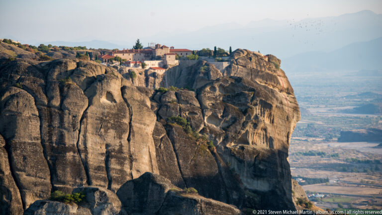 Low monastery complex lines a broad summit above a deeply grooved sandstone cliff and distant plain.
