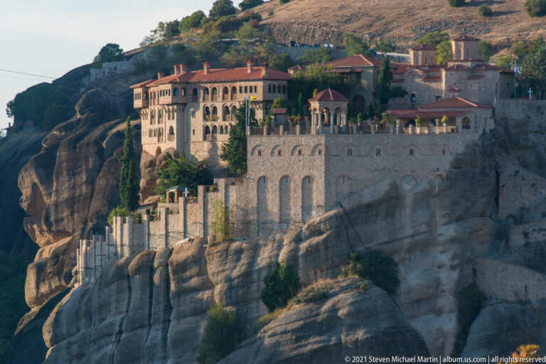 Large cliff-side monastery with repeated arched galleries, red roofs, terraces, and high stone retaining walls.