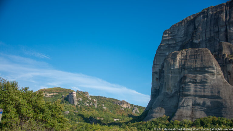 Huge vertical sandstone cliff at right overlooking a wooded valley and lower rocky ridge beneath blue sky.