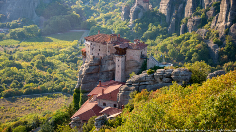 Multi-level stone monastery stands on a narrow sandstone pillar above red-roofed buildings and dense forest.