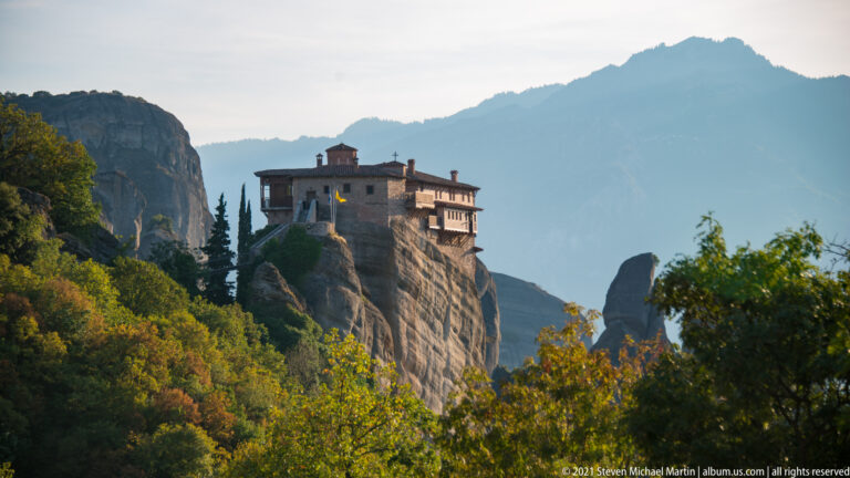 Compact stone monastery with projecting upper levels crowns a narrow sandstone column above autumn-green woodland.
