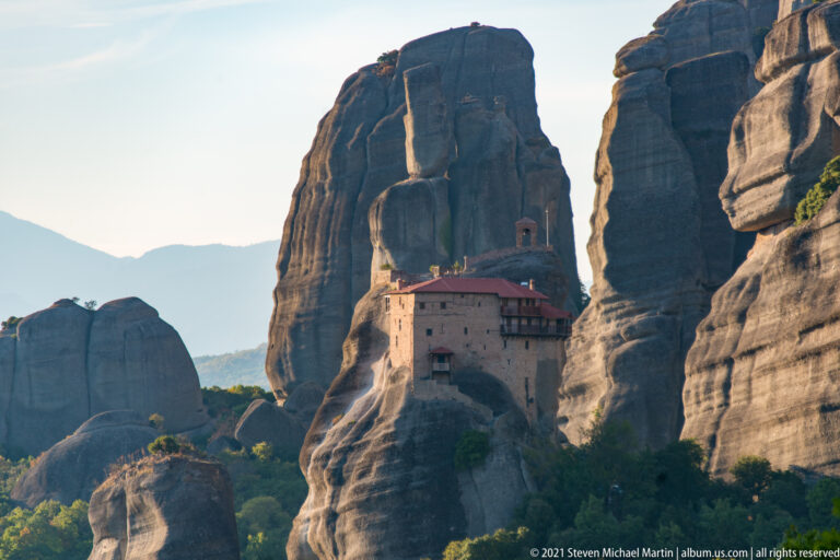Red-roofed stone monastery sits tightly against a tall sandstone tower between massive rounded cliffs.