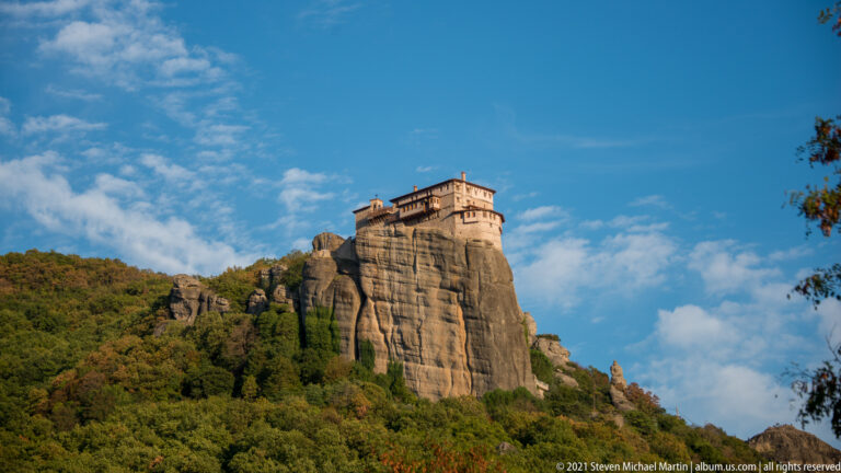 Monastery crowns a tall sandstone outcrop above wooded slopes beneath a bright blue, cloud-dappled sky.