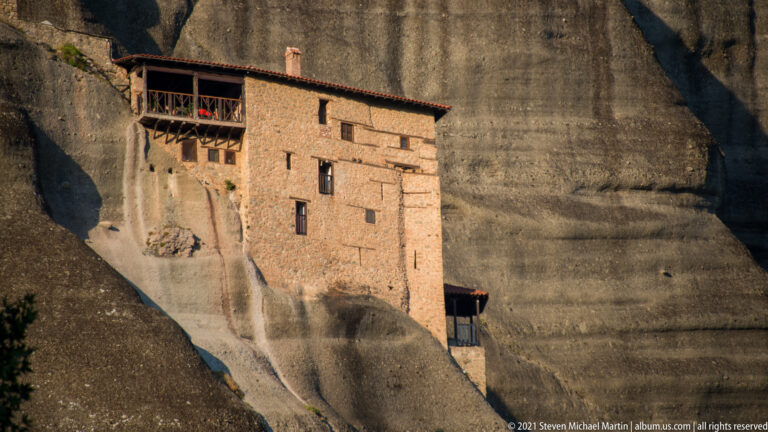 Tall stone monastic building with narrow windows and wooden galleries is built directly against a sheer sandstone wall.