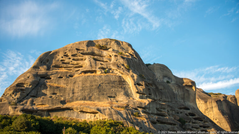 Sunlit sandstone cliff displays numerous shallow cavities, ledges, grooves, and rounded weathering forms beneath blue sky.