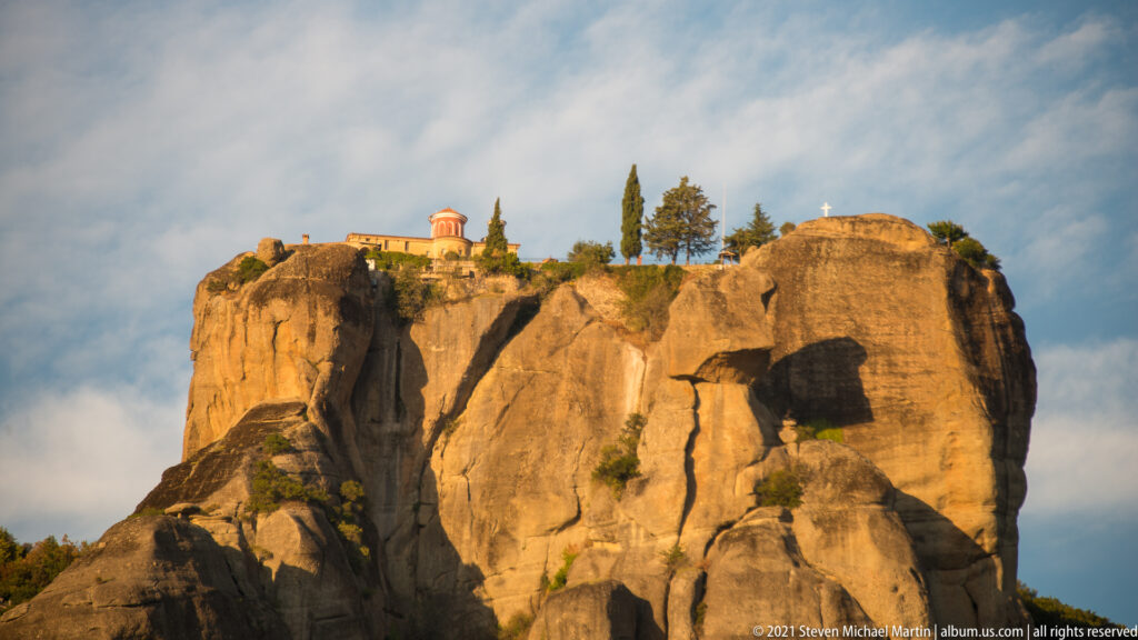 Small red-roofed monastery and cypress trees crown a massive sunlit sandstone formation with deep clefts.