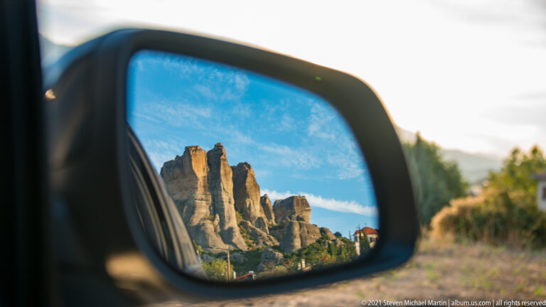 Sandstone pinnacles and blue sky reflected in a car side mirror, with the roadside blurred beyond.