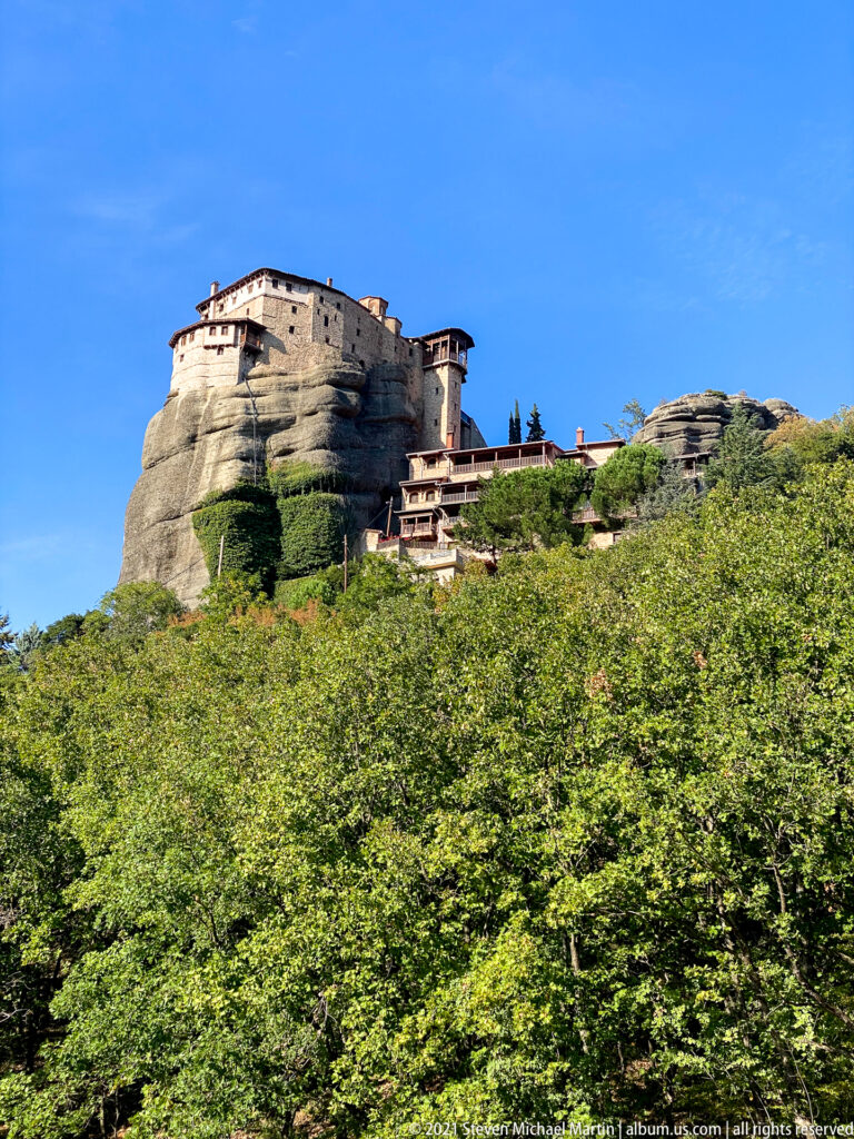 Large stone monastery with red roofs crowns a rounded sandstone summit above dense green forest.