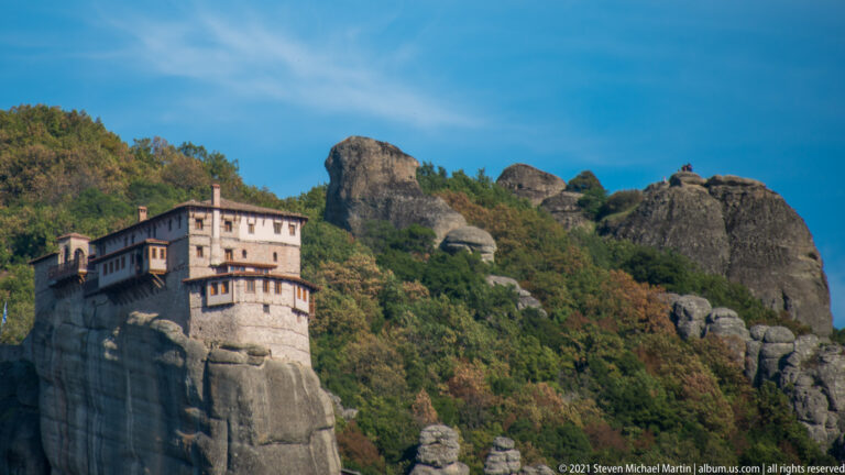 Multi-level monastery with projecting upper stories built on the top of a steep gray sandstone pillar amid forest.