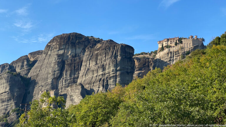 Massive sandstone wall fills the foreground left while a monastery crowns a separate rock summit beyond trees.