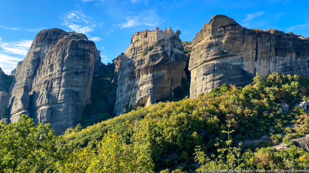 Monastery crowns the central of three towering sandstone formations above sunlit green and autumn foliage.