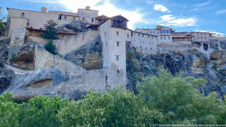 Multi-level stone monastery buildings, retaining walls, stairs, and wooden galleries rise directly from exposed sandstone above green trees.