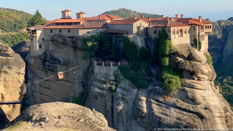 Large stone monastery with red tiled roofs and an arcaded façade crowns a sheer sandstone cliff above a deep ravine.