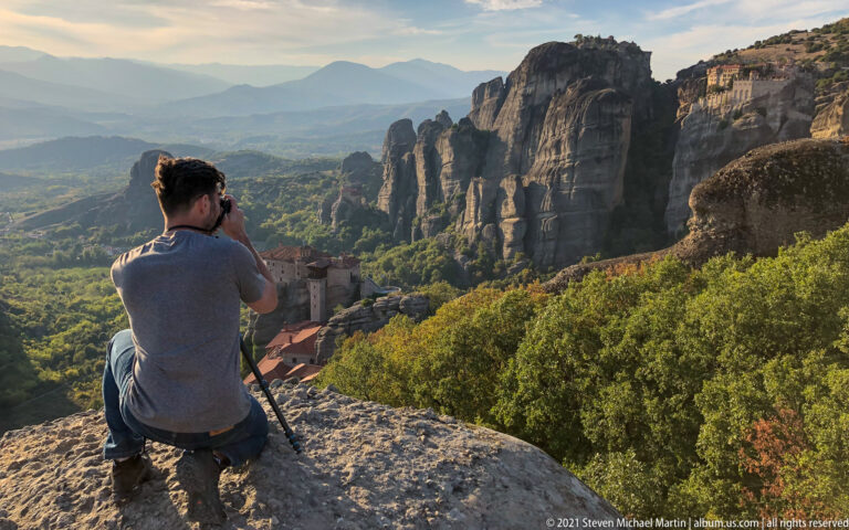 Seated visitor photographs sandstone towers and a wooded valley from a rocky overlook, with red-roofed buildings below.