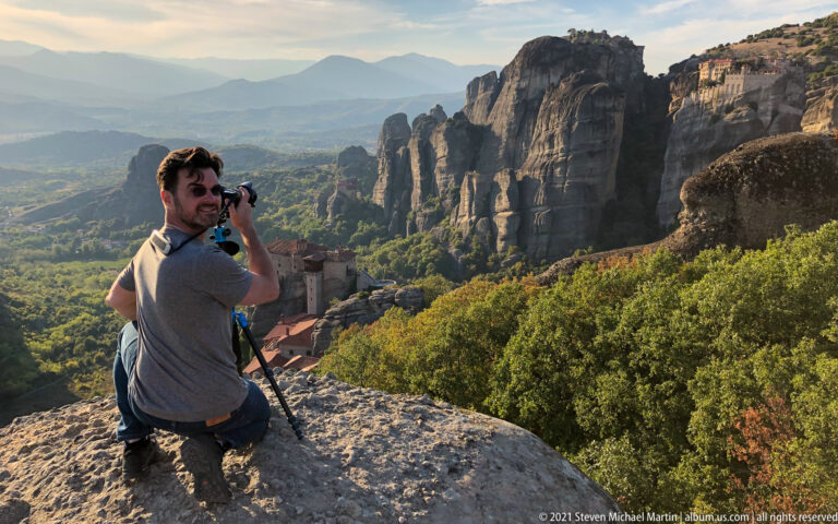 Seated visitor holding a camera turns toward the viewer above a green valley and towering sandstone formations.