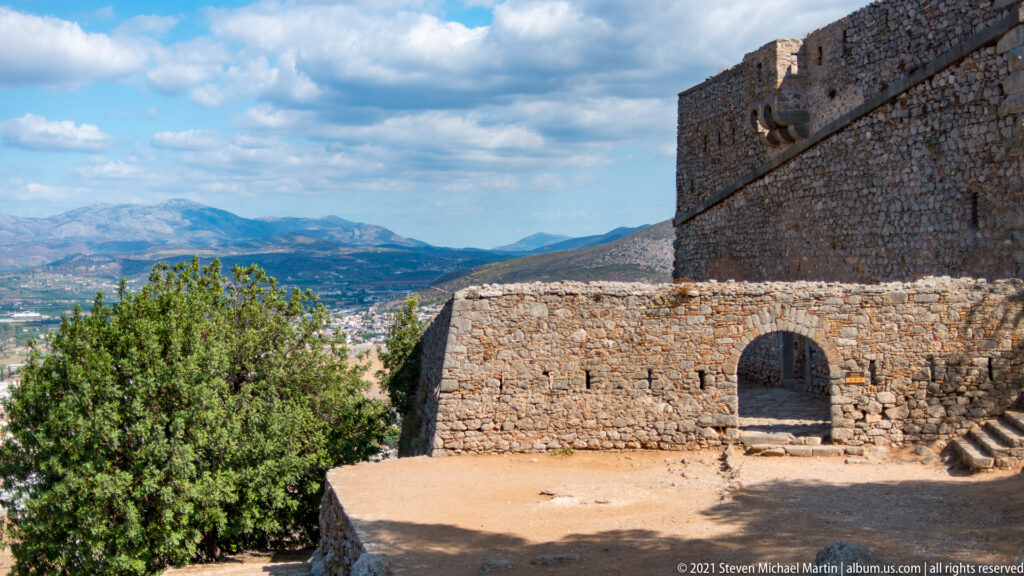 Arched gateway in a lower stone enclosure beneath a taller Palamidi wall, with mountains beyond.