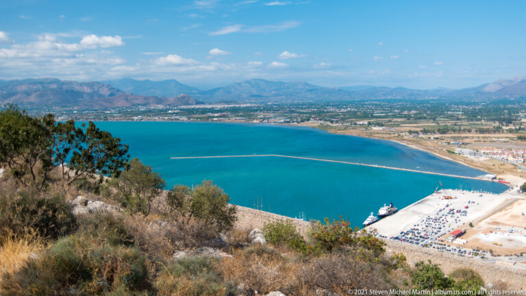 High view from Palamidi over Nafplio’s waterfront, blue bay, shoreline, and distant mountains.