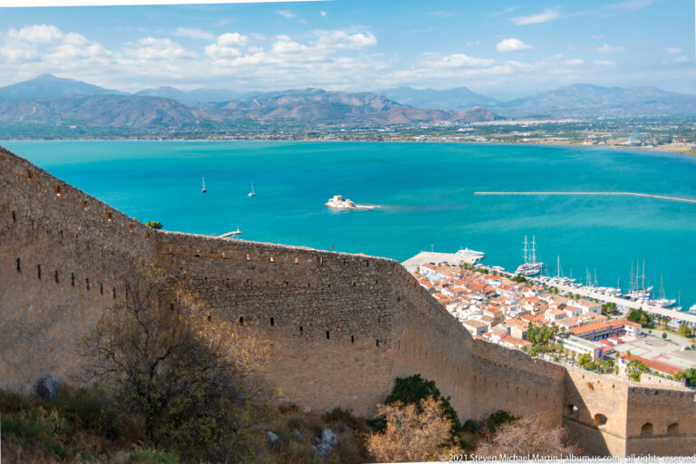 Long stone rampart descending above Nafplio, with the harbor, Argolic Gulf, and small Bourtzi island fortress beyond.