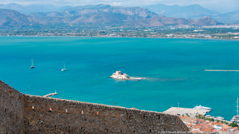 Palamidi stone rampart overlooking turquoise Nafplio harbor, with Bourtzi, sailboats, and mountains in the distance.