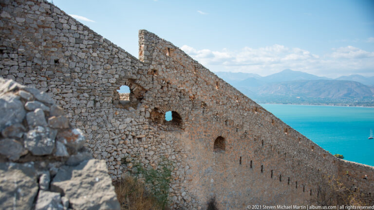 Rough stone rampart with small arched openings descending toward the bright blue Argolic Gulf.
