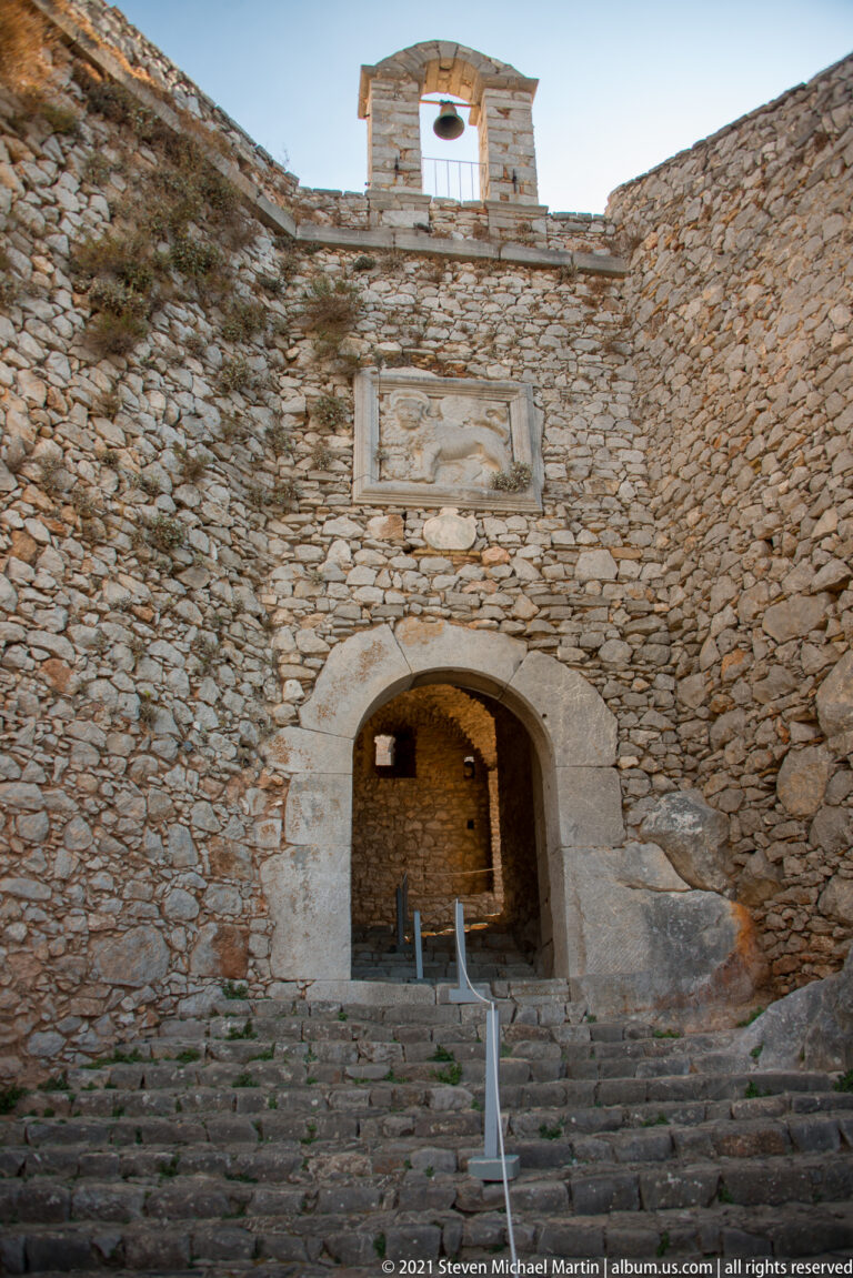 Stone steps leading through a broad fortress arch, with a small bell hanging in an opening above.