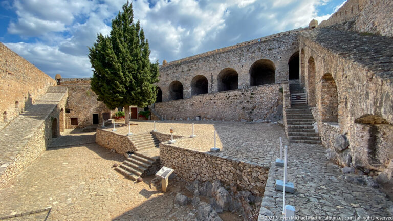 Terraced stone courtyard with steps, retaining walls, a tall cypress, and an arcaded gallery at Palamidi Fortress.